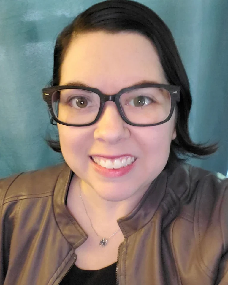 Sam Shump smiling at the camera wearing rectangular glasses and a brown leather jacket